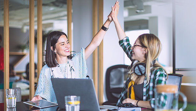 Zwei junge Frauen geben sich ein High-Five am Tisch mit einem Laptop und Tablet darauf. 