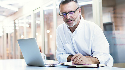 Mann mit Brille sitzt am Tisch mit Laptop und Notizbuch.