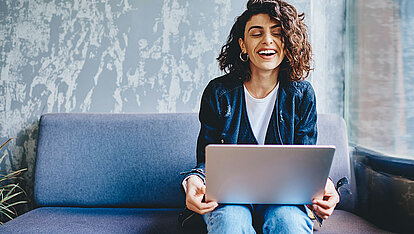 Eine Frau mit lockigem, braunem Haar sitzt auf dem Sofa und hat einen Laptop auf ihrem Schoß. Sie lacht.