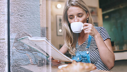 Eine Frau sitzt in einem Café am Fenster, trinkt aus einer Tasse und ließt nebenbei Zeitung. Sie trägt eine Latzhose.