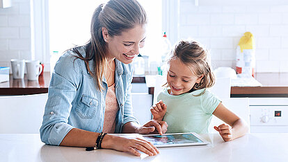 Mutter und Tochter sitzen am Küchentresen und schauen auf ein Tablet.