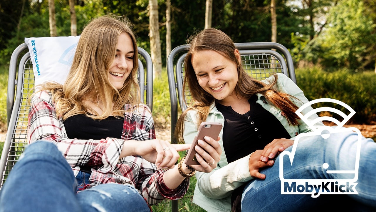 Zwei junge Frauen sitzen auf Liegestühlen. Die eine zeigt der anderen etwas auf Ihrem Handy, beide lächeln. In der rechten unteren Ecke ist ein MobyKlick-Logo in weiß abgebildet.